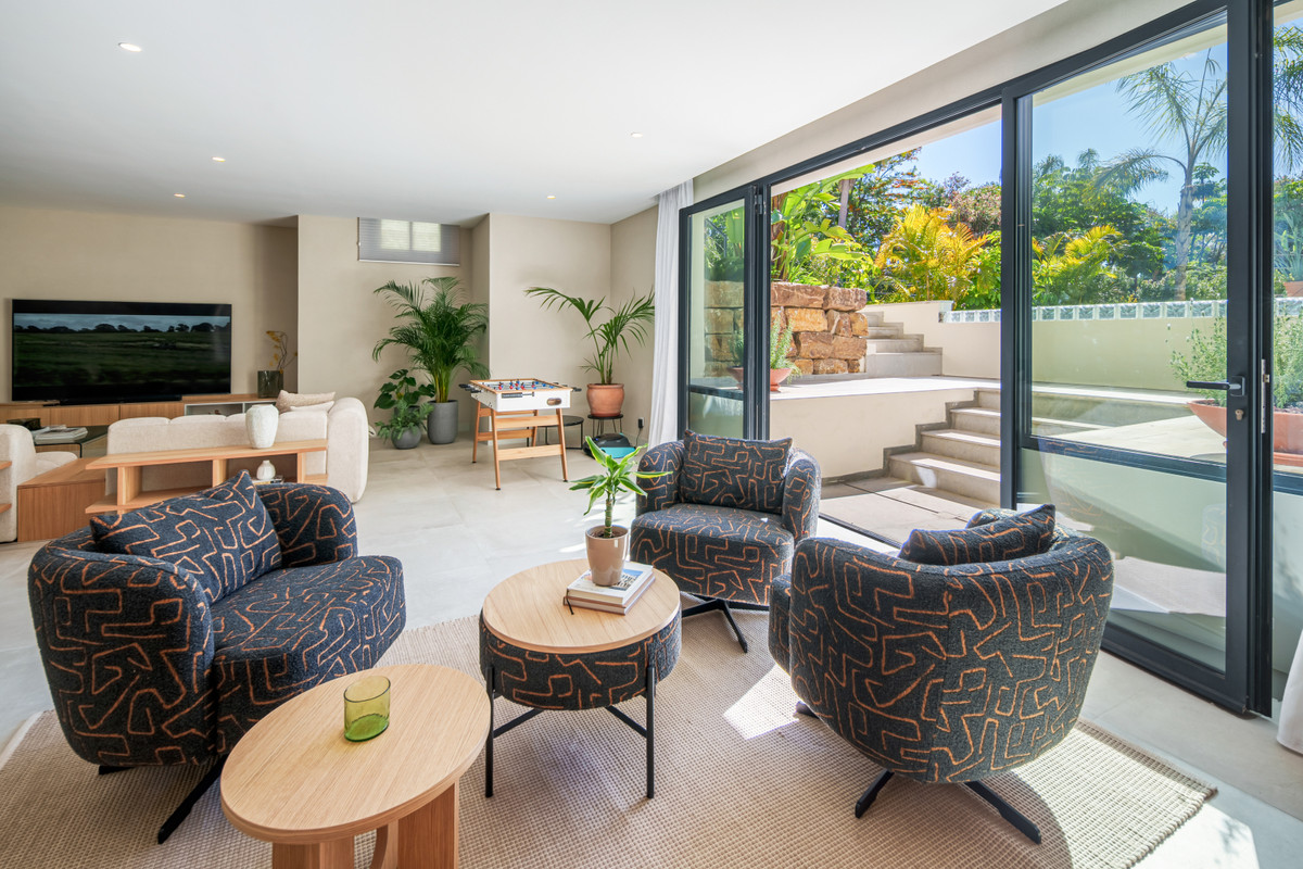 Belfry 1 – Estepona - bright open-plan living area with patterned armchairs, wooden coffee tables, and sliding glass doors opening to a lush garden terrace